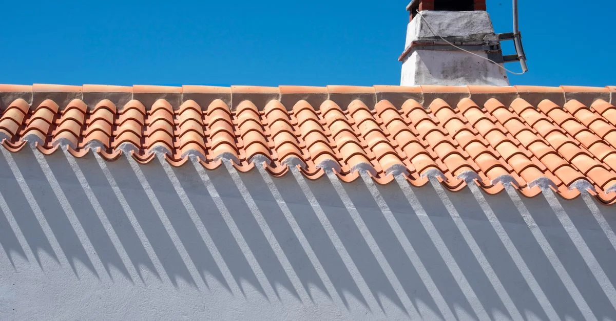 Des tuiles orange et une cheminée projettent de longues ombres sous un ciel d'un bleu éclatant.