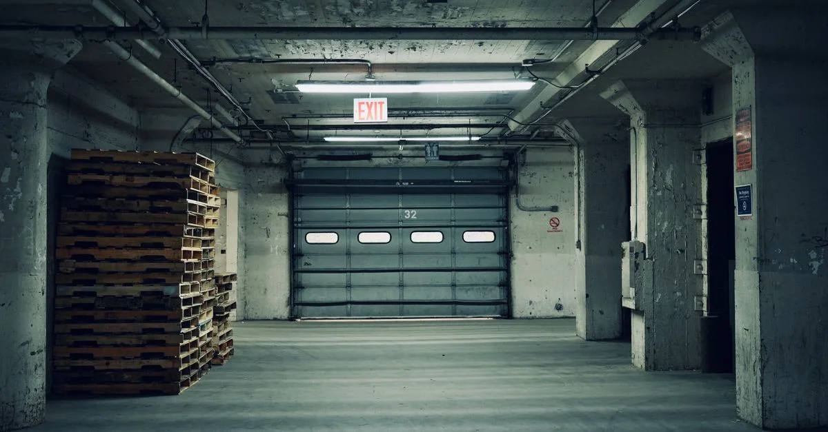 Entrepôt industriel vide avec des palettes empilées et une porte de garage fermée sous un éclairage fluorescent.