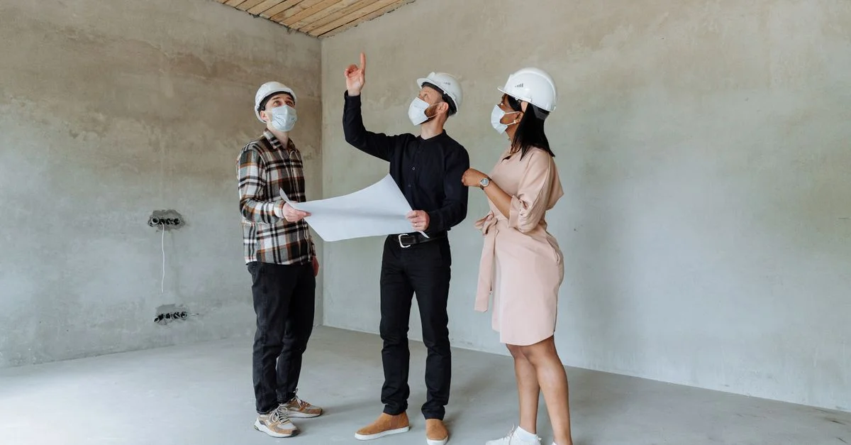 Trois architectes portant des masques et des casques discutent de plans dans un chantier intérieur.