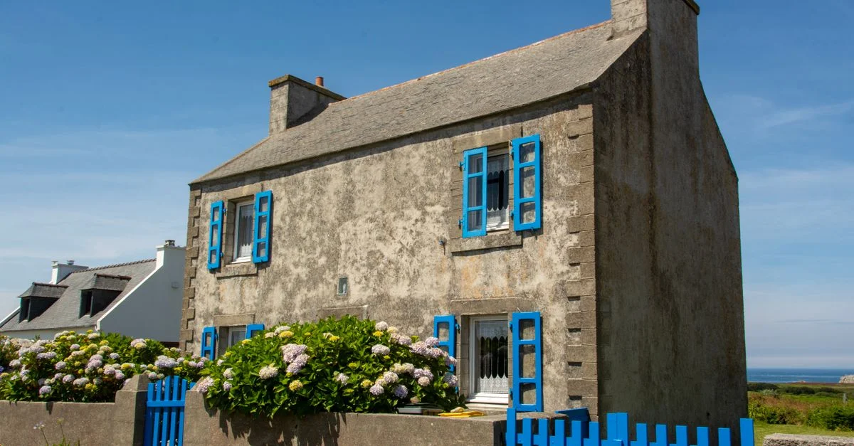 Maison rustique aux volets bleus vibrants et aux fleurs en Bretagne, parfaite pour l'inspiration de voyage.