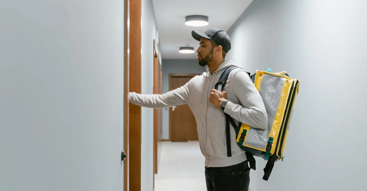 Un coursier portant un sac à dos livrant de la nourriture à une porte d'appartement à l'intérieur.