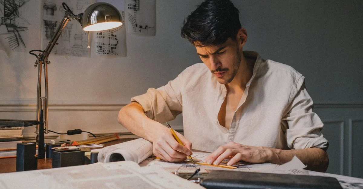 Un architecte concentré esquissant des designs à un bureau, éclairé par une lampe à l'intérieur.